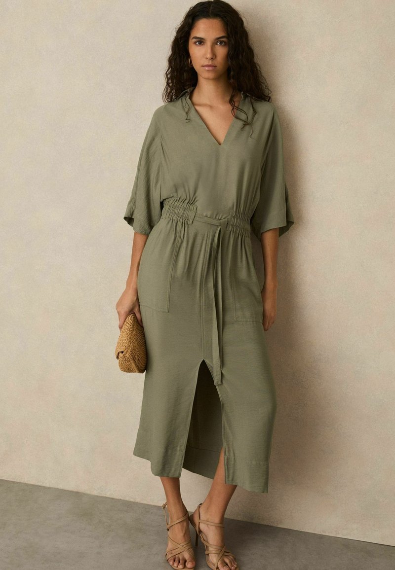 Olijfgroen midi-jurk met een V-hals, korte mouwen, elastische tailleband, voorknip en twee zakken. Gecombineerd met een gevlochten clutch en sandalen.