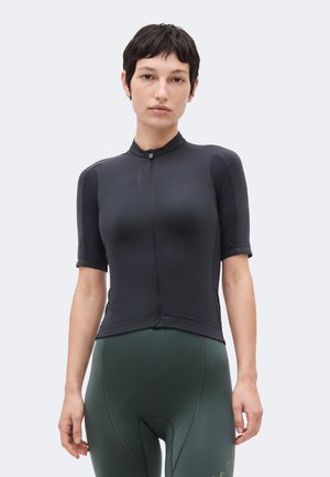 Femeie cu păr scurt și închis la culoare, purtând un tricou de ciclism negru, mulat, cu mâneci scurte și pantaloni scurți de ciclism căptușiți, de culoare verde închis.