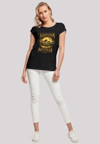 Camiseta negra de manga corta con un diseño gráfico amarillo que incluye el texto "Hakuna Matata". Combinada con pantalones blancos ajustados y zapatillas blancas.