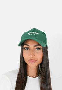 Frau mit langen dunklen Haaren, die eine grüne Sixth June Paris-Kappe und ein weißes Oberteil trägt, schaut geradeaus mit neutralem Ausdruck.