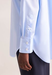 Camicia azzurra chiara con una texture liscia, con orlo arrotondato, polsini con bottoni e tre bottoni su ciascun polsino.