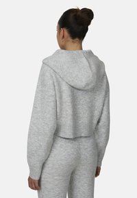 Przycięta szara bluza z kapturem o miękkiej fakturze, z szerokim kapturem oraz ściągaczami przy mankietach i u dołu. Noszona z dopasowanymi szarymi spodniami, prezentującymi luźny krój.