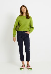 Mujer de pie con un suéter verde, pantalones recortados azul marino, zapatillas multicolores y un bolso pequeño de hombro verde oliva.