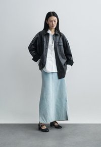 Kvinde stående iført en sort oversized læderjakke, hvid skjorte med knapper, langt lyst denimskørt med sideslids og sorte hælede sko.