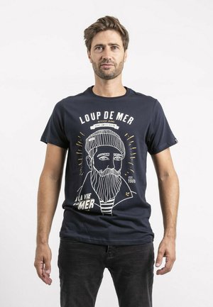 T-shirt en coton bleu marine avec un graphique blanc représentant un homme barbu portant un bonnet de marin, avec le texte "LOUP DE MER" et "À LA VIE MER" en dessous.