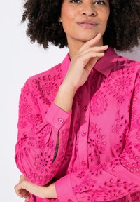 Blusa rosa ricamata con motivi floreali, dotata di chiusura a bottoni, maniche lunghe e colletto classico. Dettaglio del tessuto testurizzato visibile.