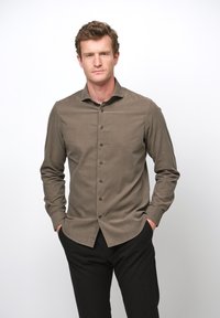 Brązowa koszula zapinana na guziki, wykonana z teksturowanej tkaniny, o wąskim kroju, z kołnierzykiem w kształcie litery V i długimi rękawami. Noszona z czarnymi spodniami.