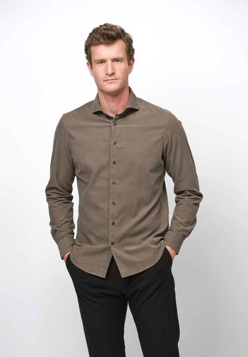 Brązowa koszula zapinana na guziki, wykonana z teksturowanej tkaniny, o wąskim kroju, z kołnierzykiem w kształcie litery V i długimi rękawami. Noszona z czarnymi spodniami.