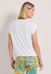 Weißes Kurzarmoberteil mit rundem Ausschnitt, kombiniert mit gemusterten mehrfarbigen Hosen, die geometrische Formen und fließende Linien aufweisen.