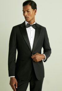 Tuxedo negru cu rever din satin; cămașă albă cu butoane; papion negru; țesătură netedă, design ajustat și formă structurată, purtat de un model.