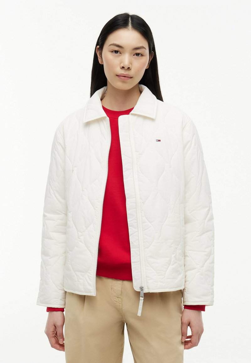 Femme portant une veste matelassée blanche par-dessus un pull rouge et un pantalon beige, debout devant un fond blanc uni.