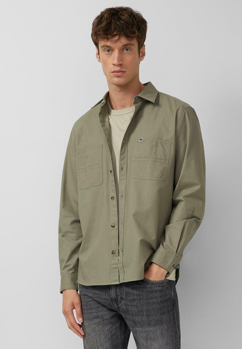Jonge man in een licht olijfgroen blouse over een beige T-shirt en donkergrijze jeans, staand met één hand in zijn zak.