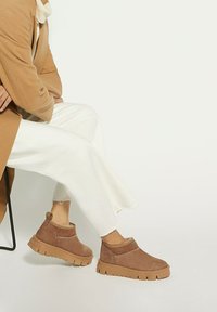 Bottines en daim marron avec une semelle épaisse et texturée et une tige basse, associées à un pantalon large crème et un manteau beige.