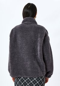 Veste en polaire gris foncé avec un col haut, surface texturée, coupe décontractée et épaules tombantes, associée à une jupe à motifs.