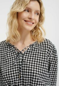 Blusa bianca e nera a motivi houndstooth con maniche lunghe, colletto arricciato e dettaglio con fiocco. Tessuto morbido con una leggera texture e rifiniture dorate.