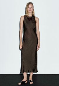 Brązowa sukienka bez rękawów o teksturowanym wykończeniu, z wysokim dekoltem. Uzupełniona ciemną naszyjnik z koralików. Nosiła czarne płaskie sandały.
