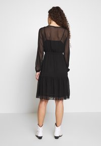 Kvinna med lockigt hår som bär en knälång svart genomskinlig klänning över en svart underklänning och vita ankelskor, stående mot en vit bakgrund vänd bort.