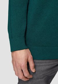 Emeraudgrüner Strickpullover mit gerippten Bündchen und Saum, dargestellt neben einer Hand, die auf einem grauen strukturierten Stoff ruht.