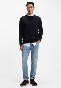 Čierny pletený sveter cez bielu pruhovanú košeľu, kombinovaný so svetlomodrými džínsami a čiernymi teniskami s hnedou podrážkou. Jednoduchý, neformálny outfit.