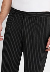 Zwarte gestreepte broeken met een getailleerde pasvorm, voorzien van zilveren streepjes, een enkele knoopsluiting en zijzakken. Geweven stof.