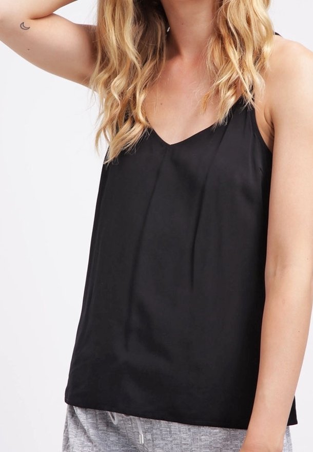 Personne blonde aux cheveux ondulés portant un haut noir sans manches, levant un bras pour toucher sa tête, pantalon gris texturé visible.
