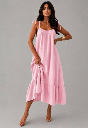 Femme aux longs cheveux foncés portant une robe maxi rose sans manches avec des bretelles nouées, tenant l'ourlet et se tenant sur un fond gris.
