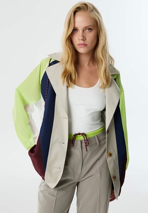 Jonge vrouw met lang blond haar draagt een kleurblok jas in beige, marineblauw, limoen en kastanjebruin over een witte top en beige broek met een touwriem.