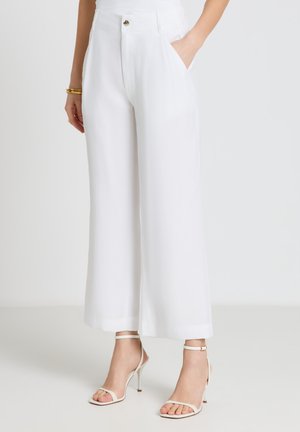 Frau trägt hoch taillierte, weit geschnittene weiße Hose mit Taschen, kombiniert mit weißen Sandalen mit hohem Absatz und Knöchelriemen sowie einem goldenen Armband.