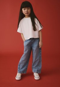 T-shirt blanc court, pantalon en denim bleu ample et baskets blanches. Fond rouge uni. L'enfant a les cheveux longs, raides et noirs.
