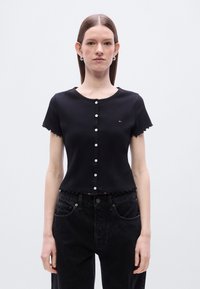 Μαύρο κοντομάνικο τοπ με κυματιστά τελειώματα, διαθέτει στρογγυλή λαιμόκοψη, κούμπωμα με κουμπιά μπροστά και μικρό λογότυπο στο στήθος.