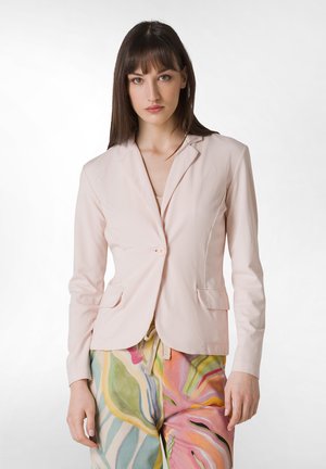 Světlý růžový dobře padnoucí blazer s jedním knoflíkem, klopami a dvěma předními kapsami, nošený přes barevně vzorované kalhoty.