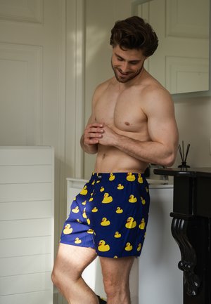 Blue boxer shorts featuring yellow rubber duck patterns, made from lightweight fabric, elastic waistband, and loose fit for comfort.