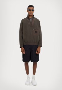Pullover verde scuro con collo a zip, dotato di tasca frontale. Abbinato a pantaloni corti neri e scarpe da ginnastica bianche, in piedi davanti a uno sfondo chiaro.