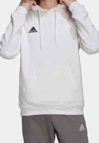 Sweat à capuche blanc avec poche avant, cordons de serrage et logo Adidas noir. Tissu doux et coupe décontractée. Associé à un pantalon gris.
