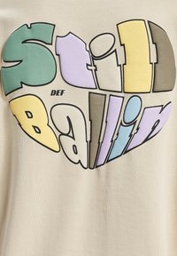 Beige T-särk, millel on südamekujuline graafika ja värviline kiri "Still Ballin' DEF" erinevates pastelltoonte ja julgete ääristustega.