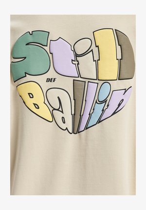 Beige T-särk, millel on südamekujuline graafika ja värviline kiri "Still Ballin' DEF" erinevates pastelltoonte ja julgete ääristustega.