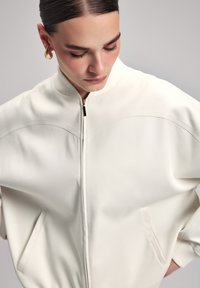Giacca bomber bianca con colletto a costine, tasche laterali e tessuto strutturato. La zip dorata aggiunge un tocco di dettaglio a un design minimalista.