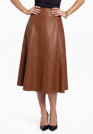 Vrouw die een bruine leren midi-rok draagt met een hoge taille, een zwart topje, zwarte puntige hakken en een gouden armband aan de linkerpols.