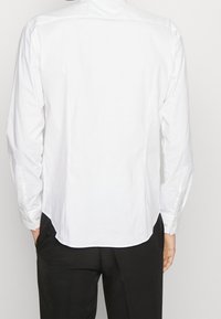 Chemise blanche à manches longues avec col boutonné, ourlet droit et tissu lisse. Vue de dos présentant un design épuré et minimaliste.