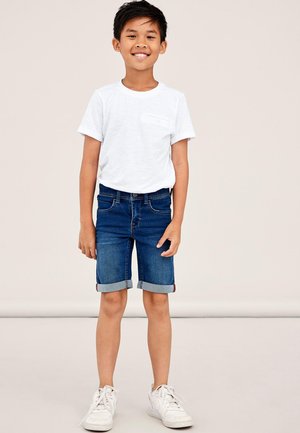 Witte t-shirt met korte mouwen en een borstzak, gecombineerd met denim shorts. De shorts hebben een opgerolde zoom. Model draagt witte sneakers.