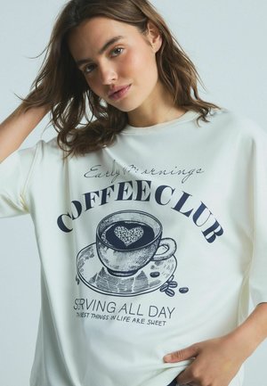 Fehér, oversized póló, amelyen egy sötétkék grafika látható egy kávéscsészéről, és a "COFFEE CLUB" valamint a "SERVING ALL DAY" feliratokkal, a csészében pedig szív mintázat található.