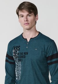 Žydrai žalios spalvos ilgomis rankovėmis marškiniai su henley apykakle, dekoruoti juodomis juostelėmis ir grafiniu tekstu skirtingais šriftai priekyje. Dviejų mygtukų užsegimas apykaklėje.