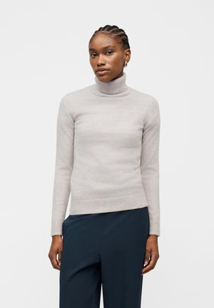 Nainen, jolla on punotut hiukset, kullanväriset renkorenkaat, vaaleanharmaa pooloneule ja tummansiniset housut, seisoo yksinkertaisen taustan edessä.