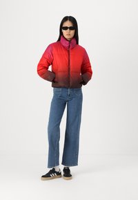 Giacca a vento rossa con gradiente scuro, dotata di colletto alto e zip frontale. Abbinata a jeans a gamba ampia blu e sneakers nere.