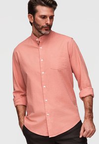 Homme portant une chemise rose à manches longues avec poignets retroussés et poche poitrine, regardant vers le bas et sur le côté.
