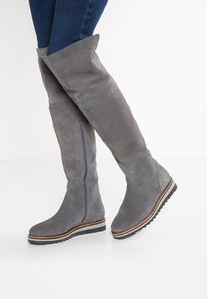 Bottes hautes en daim gris avec fermeture éclair latérale portées par-dessus un jean bleu foncé sur fond blanc uni.