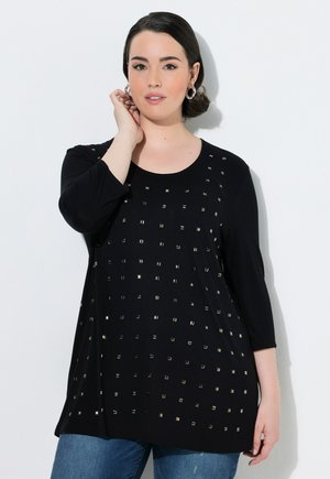 Donna con capelli scuri con una maglia nera a maniche lunghe con borchie quadrate, indossa orecchini a cerchio, in piedi davanti a uno sfondo bianco.