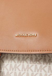 Tan leren handtas met een in goud gegraveerd "MICHAEL KORS" logo. Het interieur heeft een crèmewitte voering met een MK-patroon. Soepele textuur, slank ontwerp.