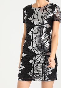 Kvinna iklädd en kortärmad, figurnära svart klänning med silverfärgade geometriska paljetter, stående mot en enkel, ljus bakgrund.