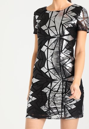 Frau in einem kurzärmligen, taillierten schwarzen Kleid mit silbernen geometrischen Paillettenmustern, die vor einem schlichten hellen Hintergrund steht.